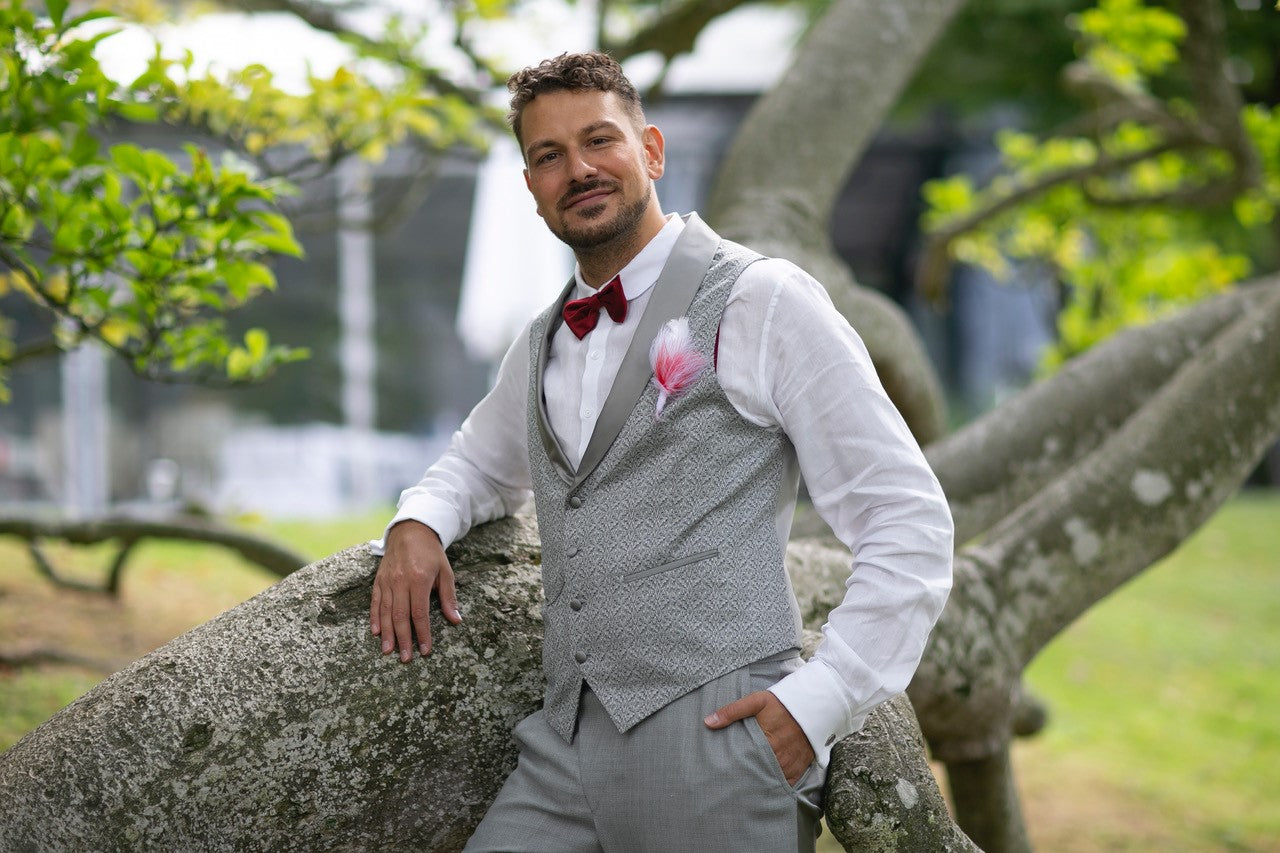 Ein Mann lehnt an einem Baum und trägt ein graues Massgilet für seine Hochzeit